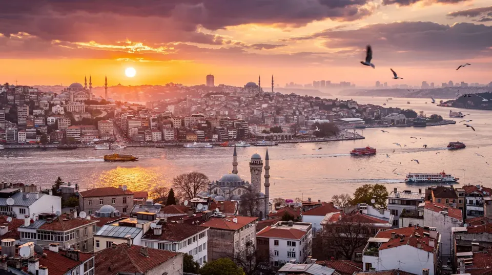 The image captures a panoramic view of Istanbul at sunset, showcasing the Bosphorus strait with historic buildings lining the waterfront and boats gently gliding on the water, creating a picturesque scene of the city’s beauty. The warm hues of the sunset reflect on the water, enhancing the overall harmony of this vibrant landscape.